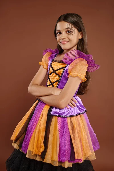 Happy kid in Halloween costume with spiderweb makeup standing with folded arms on brown backdrop — Stock Photo