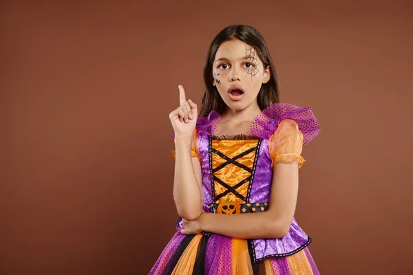 Amazed girl in Halloween costume with spiderweb makeup having idea, pointing up on brown backdrop — Stock Photo