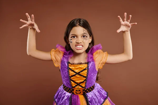 Gruseliges Kind im Halloween-Kostüm mit Spinnennetz-Make-up knurrt und gestikuliert vor braunem Hintergrund — Stockfoto