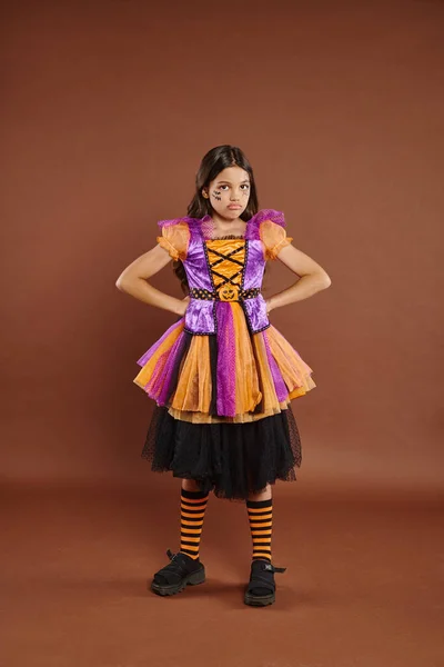 Missmutiges Mädchen im Halloween-Kostüm, das mit den Händen auf Hüften vor braunem Hintergrund steht, 31. Oktober — Stockfoto