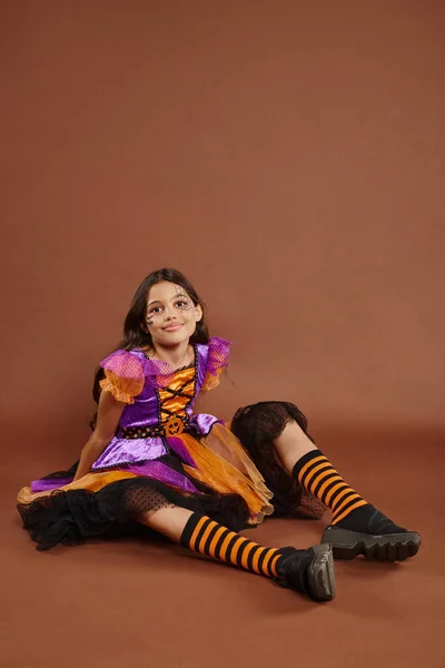 Cheerful girl in colorful Halloween dress costume sitting and looking at camera on brown backdrop — Stock Photo
