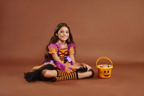 Cheerful girl in colorful Halloween costume sitting near bucket with candies on brown backdrop — Stock Photo