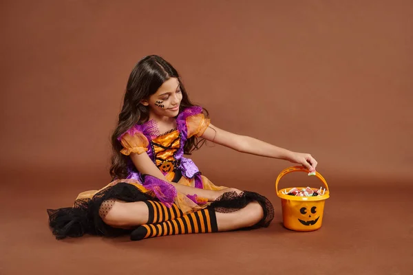 Menina positiva no traje de Halloween colorido sentado perto do balde com doces no fundo marrom — Fotografia de Stock