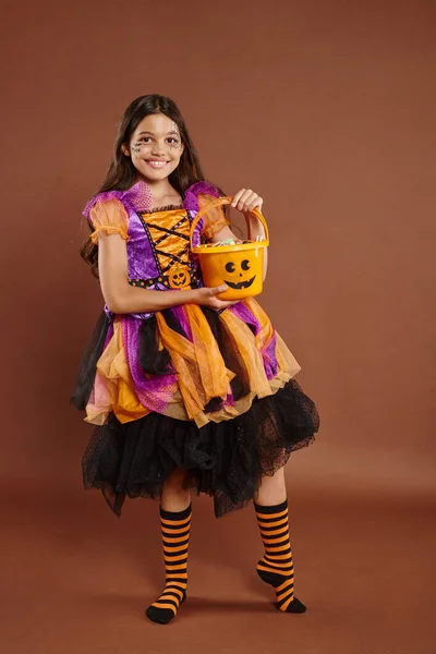 Chica feliz en colorido traje de Halloween sosteniendo cubo con caramelos y de pie sobre fondo marrón - foto de stock