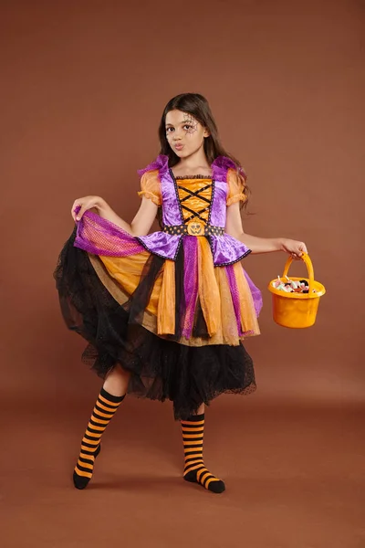 Adorable chica en traje de Halloween celebración de cubo con caramelos y la celebración de la falda en el fondo marrón - foto de stock