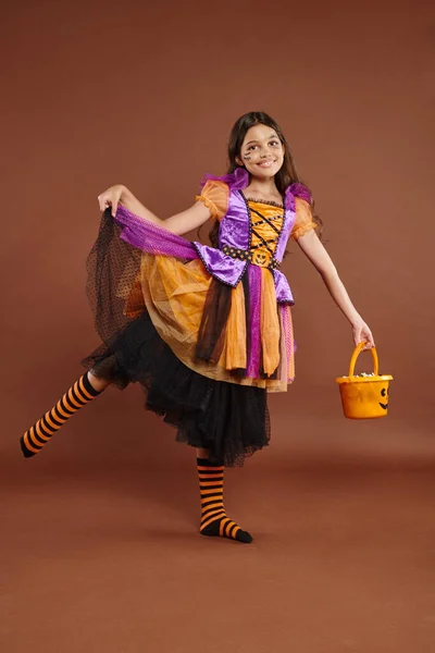 Cheerful girl in Halloween costume holding bucket with candies and holding skirt on brown backdrop — Stock Photo