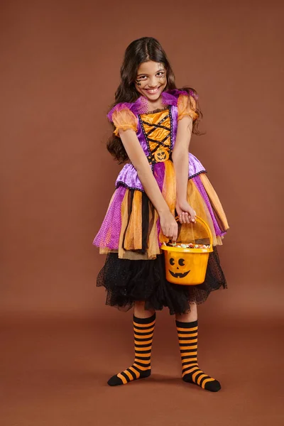 Entzückendes Mädchen im Halloween-Kostüm hält Eimer mit Bonbons und hält Rock auf braunem Hintergrund — Stockfoto