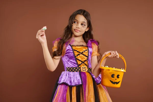 Menina bonito no traje de Halloween segurando balde e olhando para doces embrulhados no fundo marrom — Fotografia de Stock