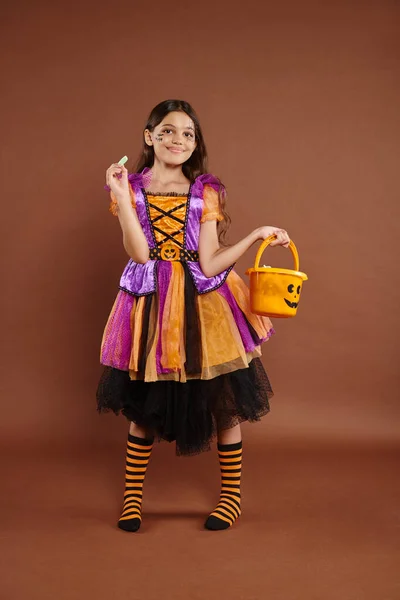 Chica encantadora en traje de Halloween sosteniendo cubo y mirando caramelos envueltos en fondo marrón - foto de stock
