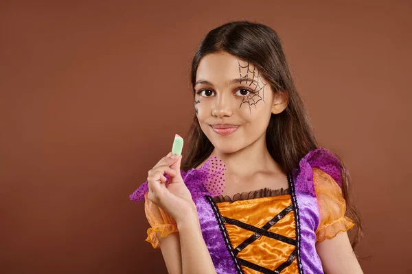 Cheerful girl in Halloween costume holding sweet jelly candy on brown backdrop, trick or treat — Stock Photo