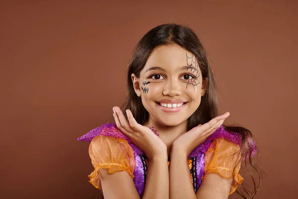 Fröhliches Mädchen im Halloween-Kostüm und Spinnennetz-Make-up gestikulierend auf braunem Hintergrund, Trick oder Leckerbissen — Stockfoto