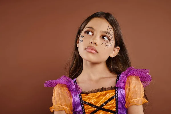 Pensive girl in colorful costume with Halloween makeup looking away on brown background, October — Stock Photo