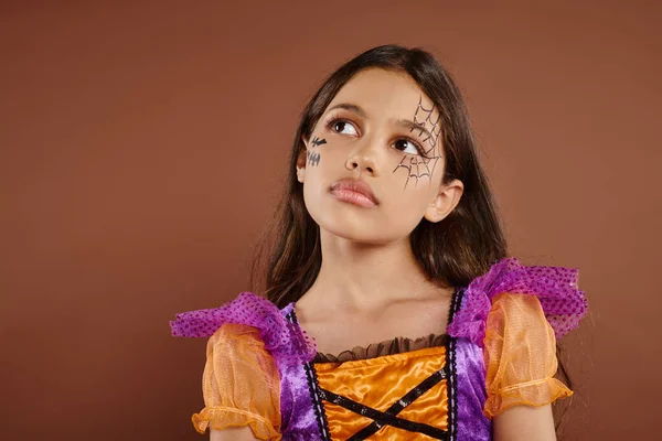 Menina pensativa em traje colorido com maquiagem Halloween olhando para longe no fundo marrom, Outubro — Fotografia de Stock