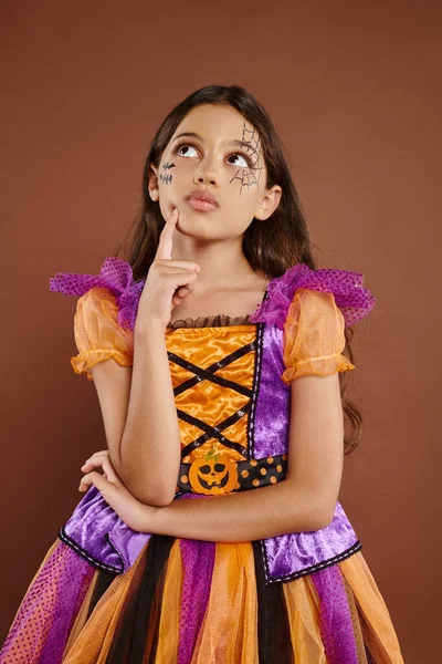 Menina pensativa em traje colorido com maquiagem Halloween olhando para longe no fundo marrom, Outubro — Fotografia de Stock