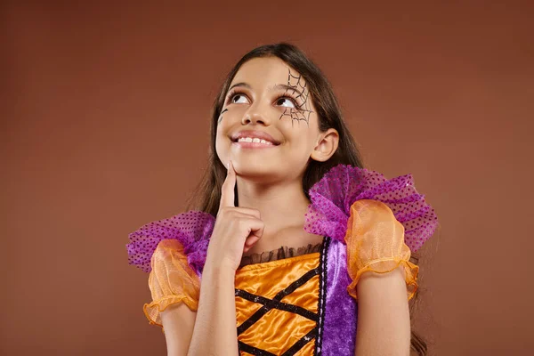 Menina sonhadora em traje colorido com maquiagem Halloween olhando para longe no fundo marrom, rosto feliz — Fotografia de Stock