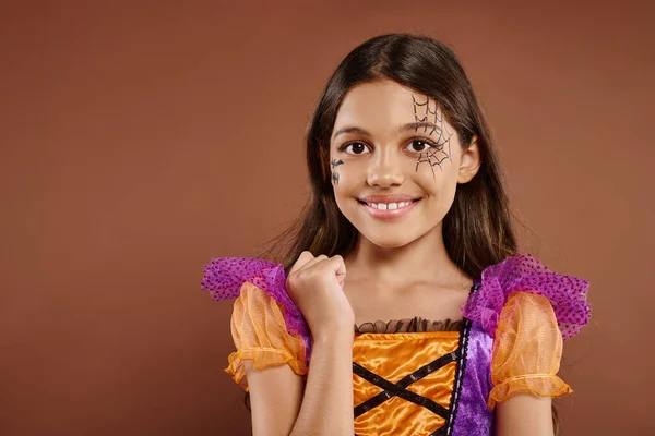 Chica en traje colorido con maquillaje de Halloween mirando a la cámara sobre fondo marrón, cara feliz - foto de stock