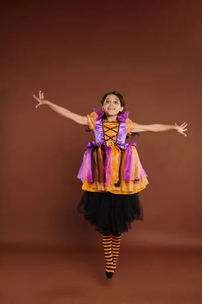 Chica feliz en colorido disfraz de Halloween mirando a la cámara y saltar sobre fondo marrón, levitar - foto de stock