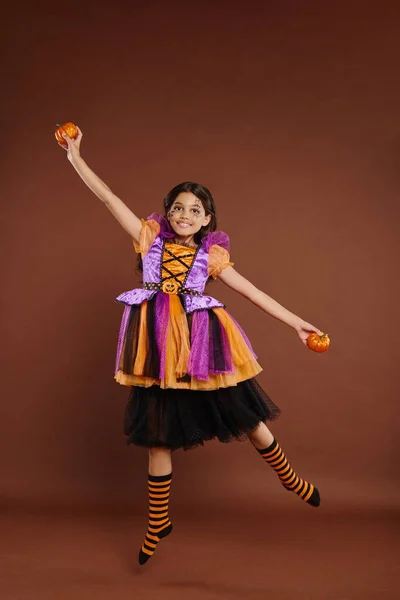 Happy girl in Halloween costume levitating with pumpkins on brown background, magic concept — Stock Photo