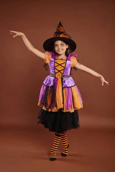 Anmutiges Mädchen im Halloween-Kostüm und Hexenhut posiert auf braunem Hintergrund, magisches Konzept — Stockfoto