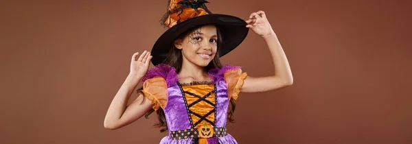 Chica feliz en disfraz de Halloween y sombrero puntiagudo posando sobre fondo marrón, pequeña bandera de bruja - foto de stock