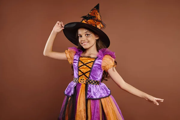 Fröhliches Mädchen im Halloween-Kostüm und Zipfelmütze posiert auf braunem Hintergrund, kleine Hexe — Stockfoto
