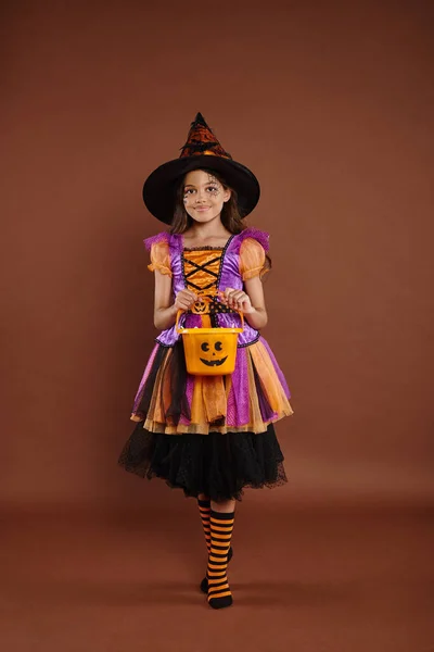 Chica feliz en disfraz de Halloween y sombrero puntiagudo de pie con balde de caramelo sobre fondo marrón - foto de stock