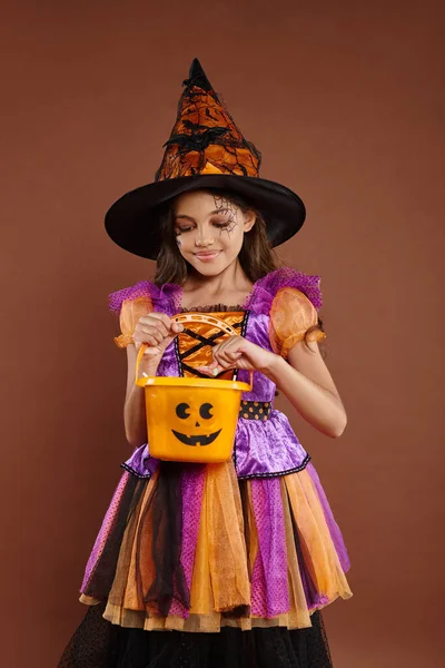 Chica alegre en disfraz de Halloween y sombrero apuntado mirando cubo de caramelo sobre fondo marrón - foto de stock