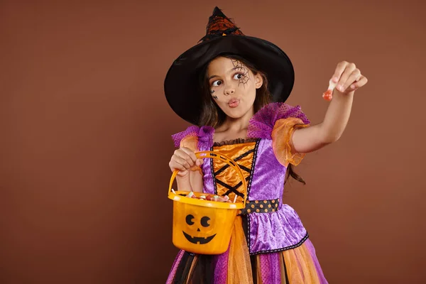 Chica divertida en disfraz de Halloween y sombrero puntiagudo sosteniendo cubo y mostrando dulces sobre fondo marrón - foto de stock