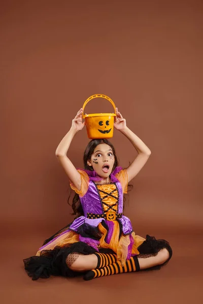 Menina surpreso no traje de Halloween segurando balde de doces na cabeça e sentado no fundo marrom — Fotografia de Stock