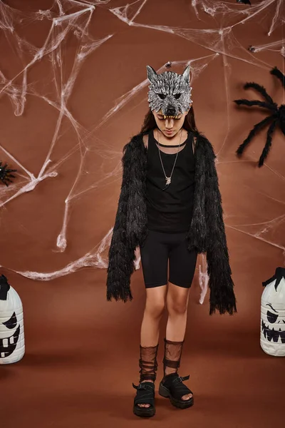 Mädchen im Werwolfkostüm mit Maske und schwarzer Kunstpelzjacke steht auf braunem Hintergrund mit Spinnwebe — Stockfoto