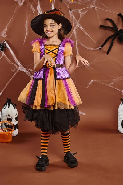Alegre chica en sombrero de bruja y vestido bailando cerca de cubo con dulces en marrón, concepto de Halloween - foto de stock