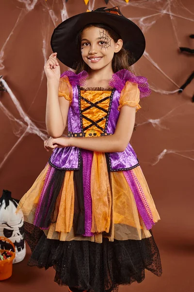 Menina positiva em traje de bruxa e chapéu pontiagudo sorrindo no fundo marrom, conceito de Halloween — Fotografia de Stock