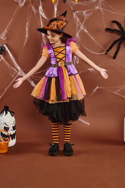 Menina positiva no traje de bruxa e apontou chapéu gesticulando perto de balde doces, conceito de Halloween — Fotografia de Stock