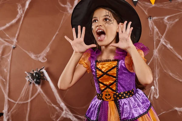 Эмоциональная девушка в шляпе ведьмы и костюме Хэллоуина кричит и жестикулирует на коричневом фоне — стоковое фото