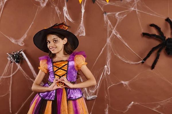 Cheerful little witch in Halloween costume and pointed hat showing heart gesture on brown backdrop — Stock Photo