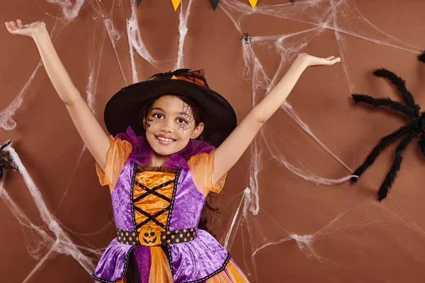Alegre brujita en disfraz de Halloween y sombrero puntiagudo con las manos levantadas sobre fondo marrón - foto de stock