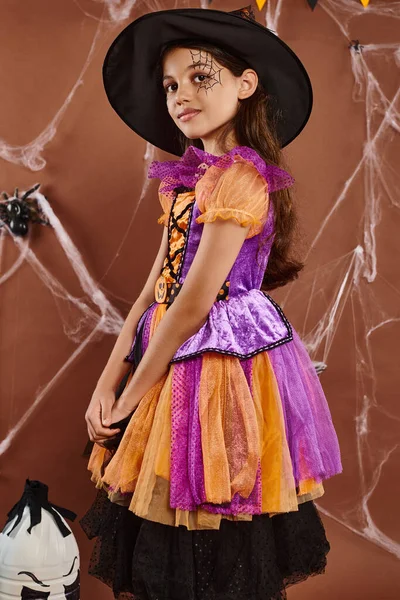 Sonriente pequeña bruja en disfraz de Halloween y sombrero puntiagudo de pie sobre fondo marrón, linda chica - foto de stock