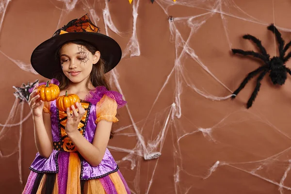 Cheerful girl in Halloween witch costume and pointed hat holding pumpkins on brown backdrop — Stock Photo