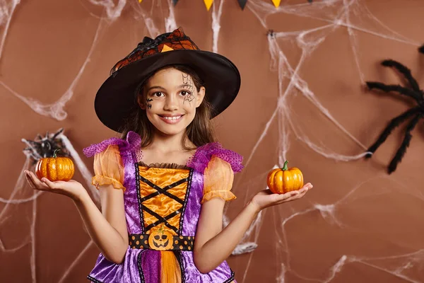 Chica alegre en disfraz de bruja de Halloween y sombrero puntiagudo posando con calabazas sobre fondo marrón - foto de stock