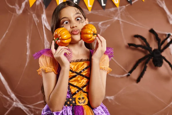 Funny girl with spiderweb makeup puffing cheeks and  holding pumpkins on brown backdrop, Halloween — Stock Photo