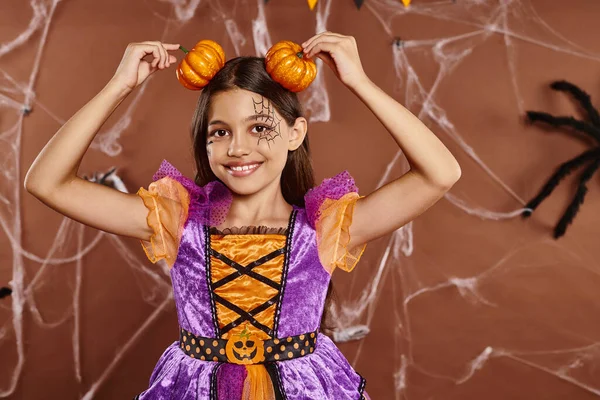 Chica divertida con maquillaje de tela de araña sonriendo y sosteniendo calabazas cerca de la cabeza en el fondo marrón, Halloween - foto de stock