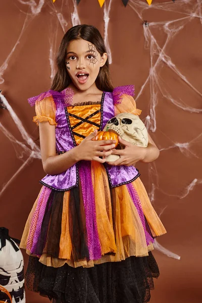 Chica sorprendida en disfraz de Halloween sosteniendo calabazas y cráneo en el fondo marrón, temporada espeluznante - foto de stock