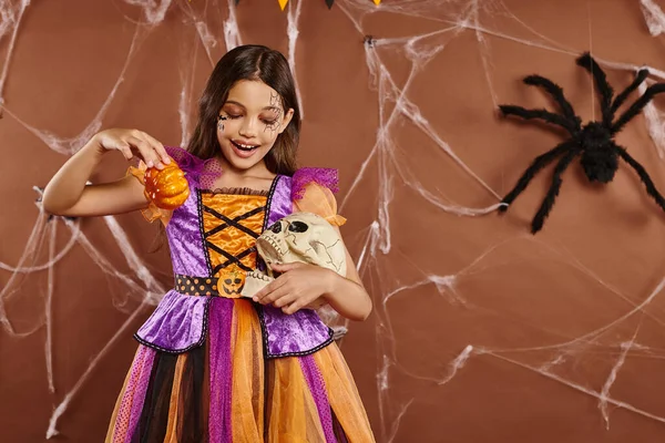 Lächelndes Mädchen im Halloween-Kostüm mit Kürbissen und Totenkopf vor braunem Hintergrund, gruselige Jahreszeit — Stockfoto