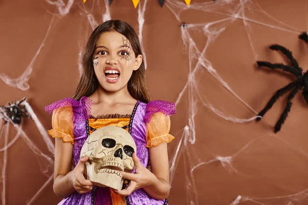 Chica en disfraz de Halloween sosteniendo el cráneo y gruñendo en la cámara sobre fondo marrón, temporada espeluznante - foto de stock