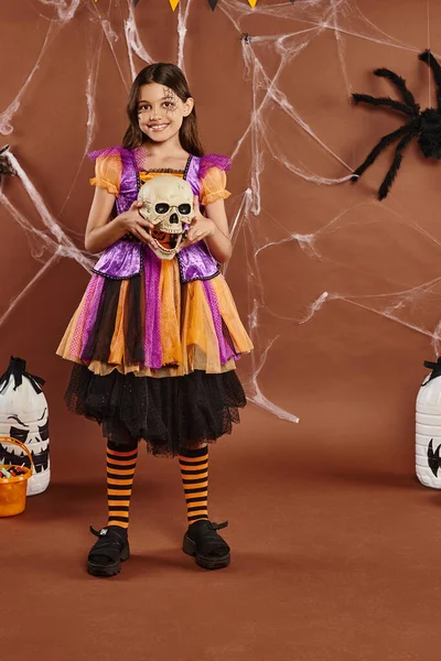 Cheerful girl in dress holding skull and smiling on brown background, Halloween spooky season — Stock Photo