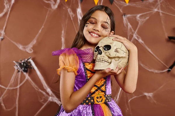 Encantadora menina no vestido segurando crânio e sorrindo no fundo marrom, Halloween estação assustadora — Fotografia de Stock