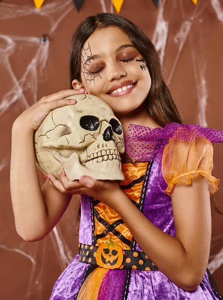 Menina positiva no vestido segurando crânio e sorrindo no fundo marrom, Halloween estação assustadora — Fotografia de Stock