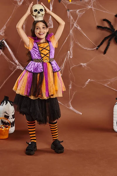 Menina alegre no vestido de Halloween de pé com crânio na cabeça no fundo marrom, estação assustadora — Fotografia de Stock