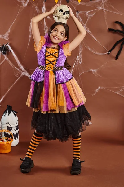 Веселая девушка в Хэллоуинском платье стоит с черепом на голове на коричневом фоне, жуткий сезон — стоковое фото
