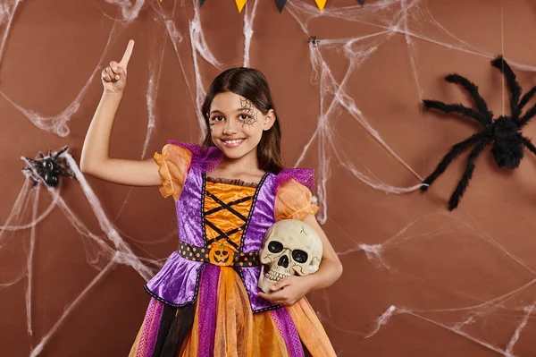 Cheerful girl in Halloween dress standing with skull and pointing up with finger on brown background — Stock Photo